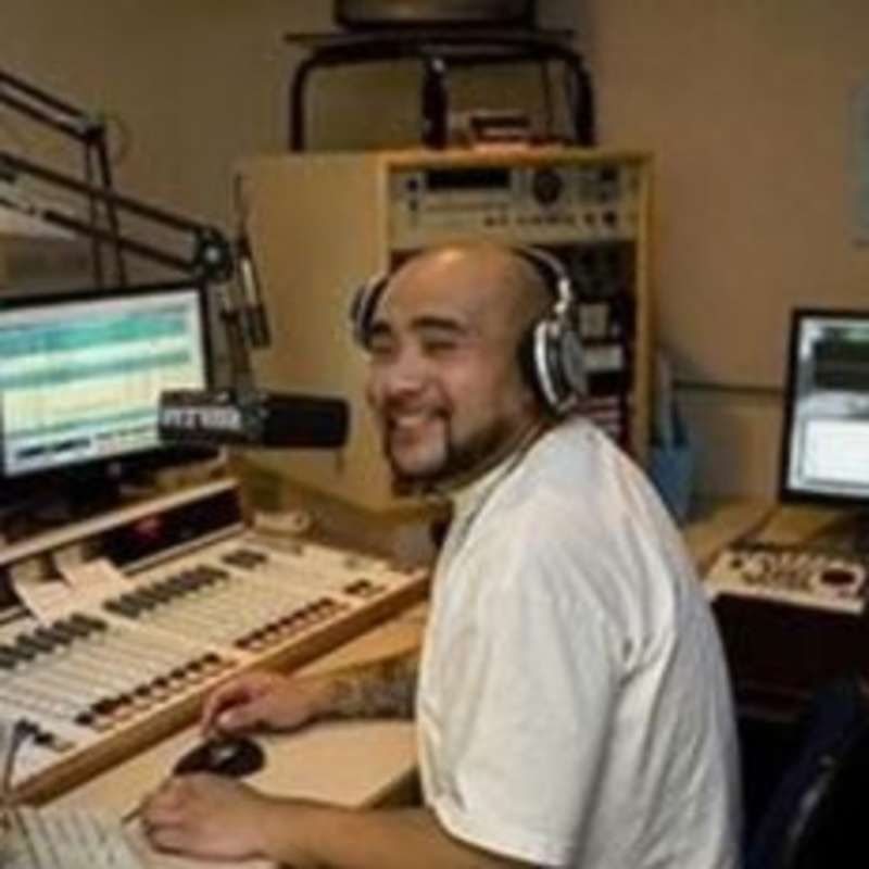 A man sitting while smiling at a desk in front of a radio, listening intently.