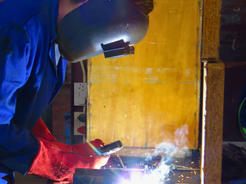 A person wearing a welding helmet and red gloves welds metal, emitting bright sparks and smoke. The scene is set against a wooden backdrop.