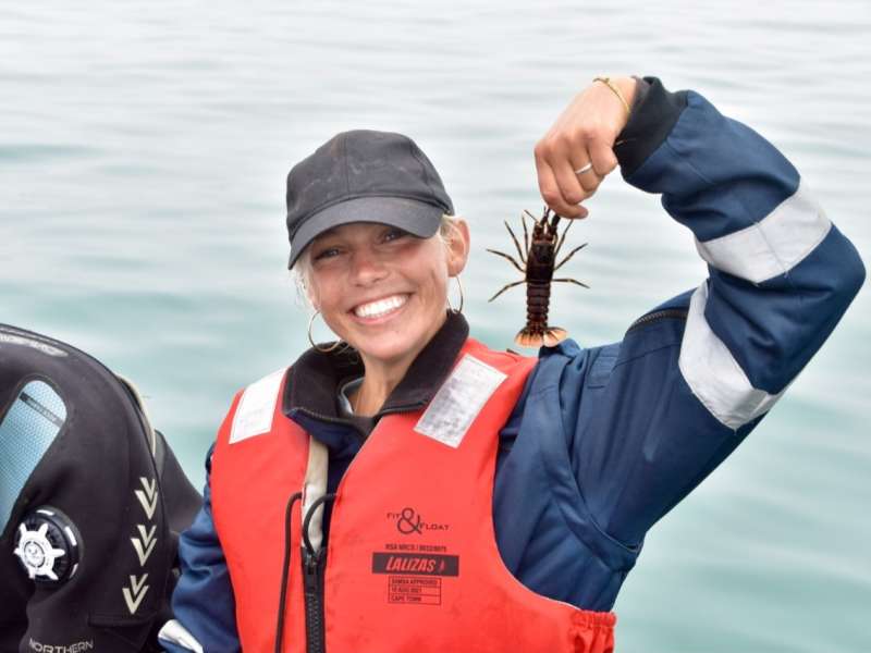 A smiling team member in a life jacket holds up a rock lobster retrieved during a crayfish monitoring survey on a research vessel off the Lüderitz coast.