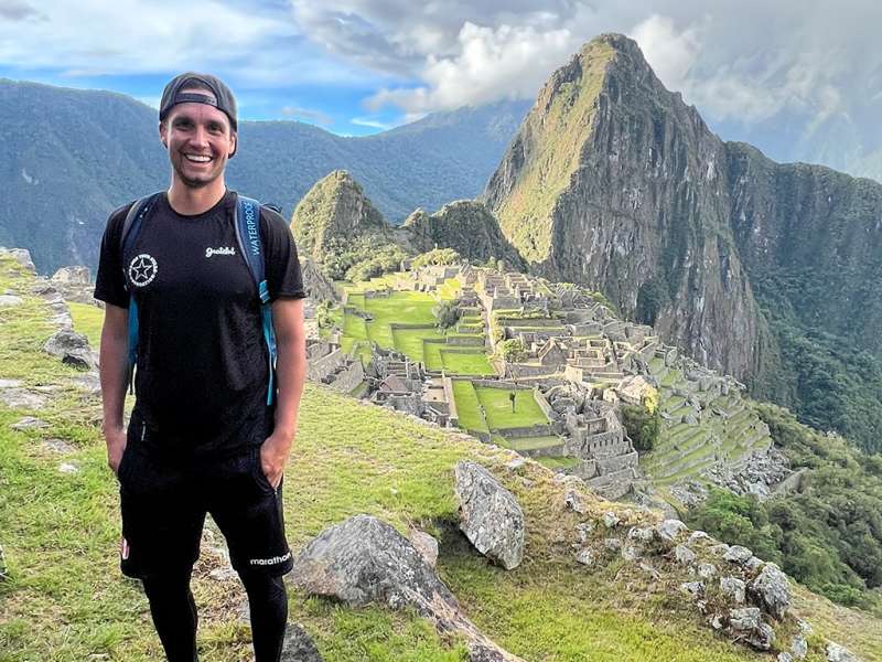 A smiling traveler stands in the foreground with the ancient Incan citadel of Machu Picchu and towering green mountains behind him.