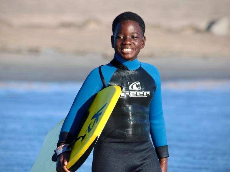 A student in a wetsuit smiles at the camera on the Lüderitz beach, holding a yellow bodyboard with the South Atlantic in the background.