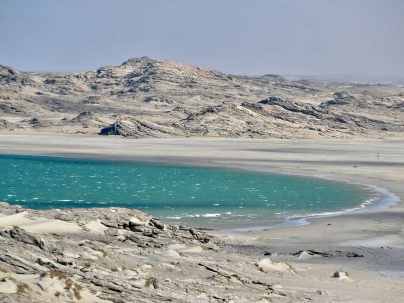 A turquoise lagoon nestled between dramatic rocky desert terrain and pale sand dunes on the Namibian coastline, under a hazy sky.