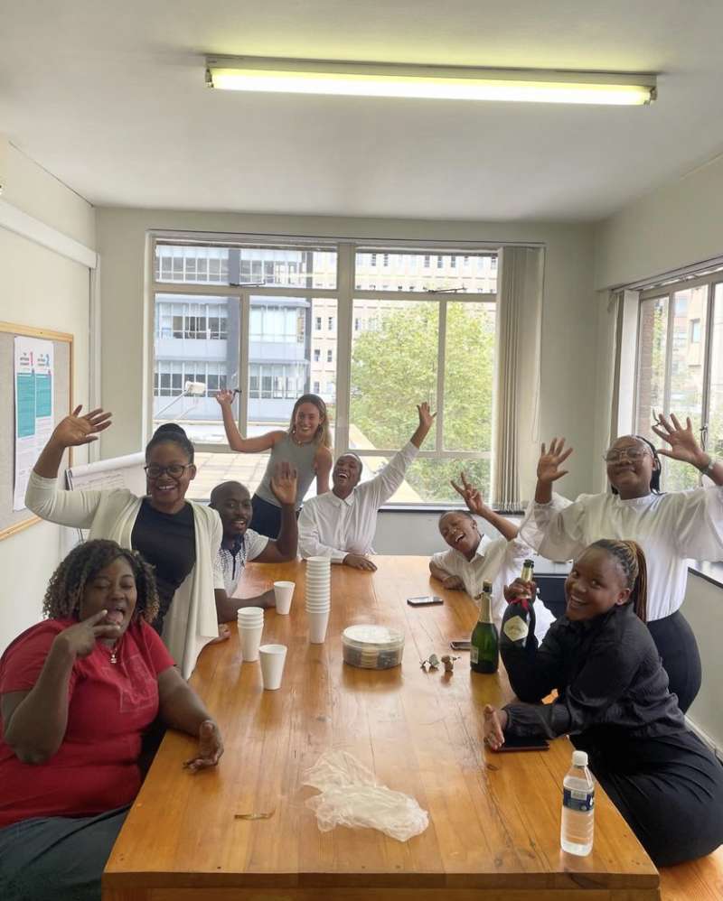 A volunteer and the team at the law clinic smile and wave at the camera, sat around a table celebrating with champagne!