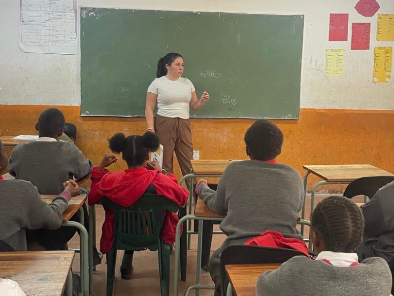 A volunteer stands in a classroom full of children, in front of a chalk board. She has her left hand out in front of her as she explains something to the children. 
