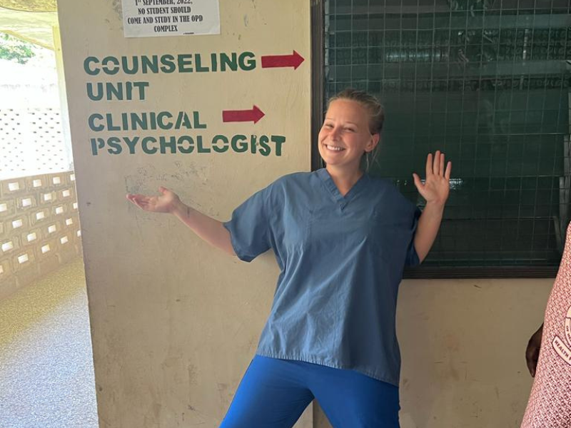 A volunteer stands in her scrubs, smiling and posing underneath a sign and arrows that read 'counseling unit' and 'clinical psychologist'. 