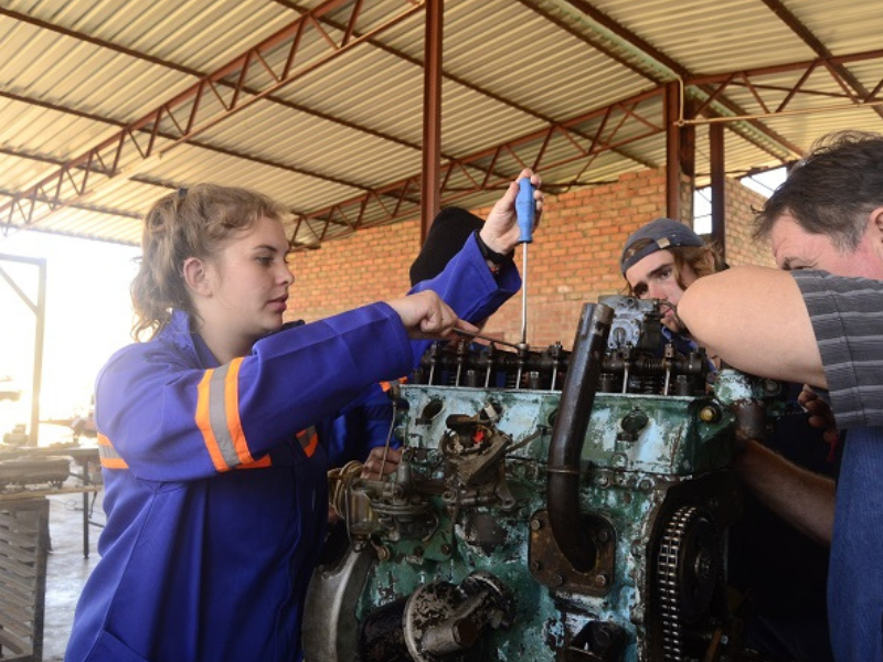 A woman in a blue work uniform with orange stripes repairs a large engine, surrounded by three men. The setting is industrial, with a focused atmosphere.