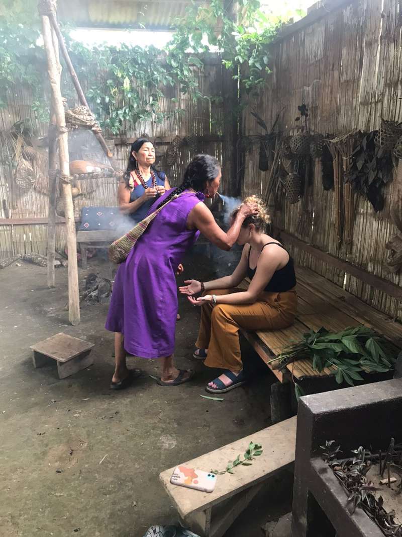 A woman in traditional clothes blows smoke on another's head. 