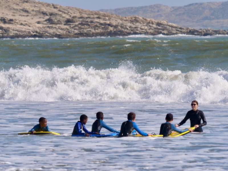 An intern in a wetsuit guides a line of primary school students on yellow bodyboards in the ocean, with crashing waves and the Lüderitz coastline behind them.