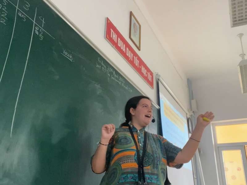 Female TEFL teacher standing in front of chalk board, wearing head microphone and gesturing with arm.