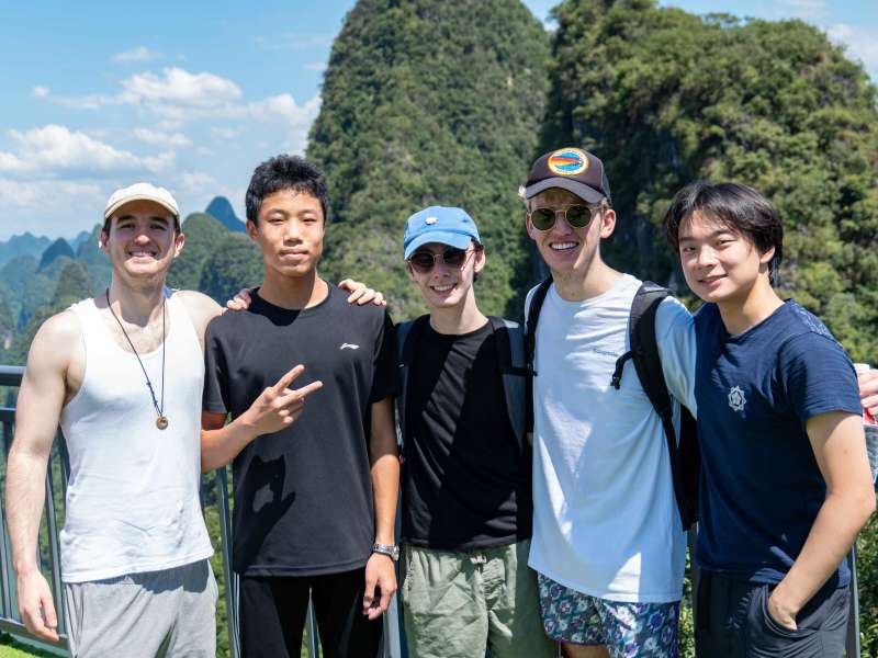 Five young men pose arm in arm at a scenic overlook above the Li River, with green karst mountains rising behind them.