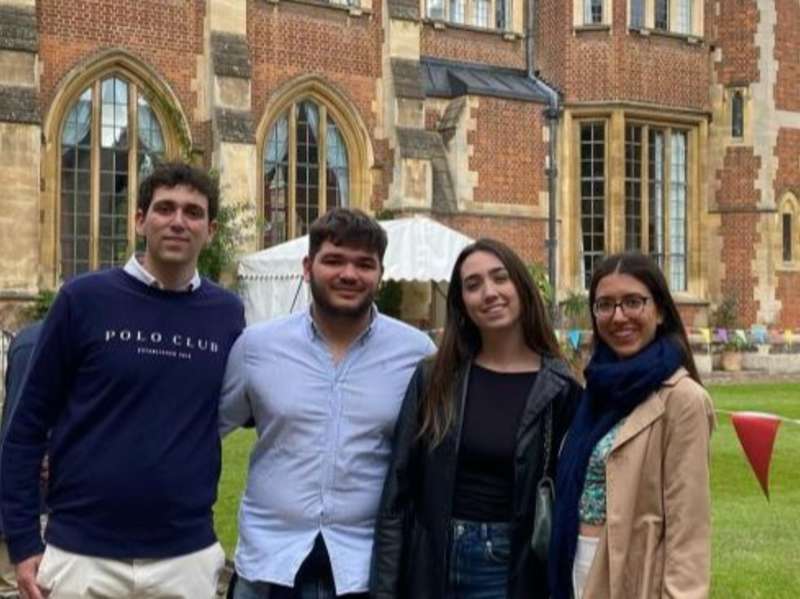 Four students standing together on a lawn in front of a historic brick university building with large arched windows in Cambridge, England.