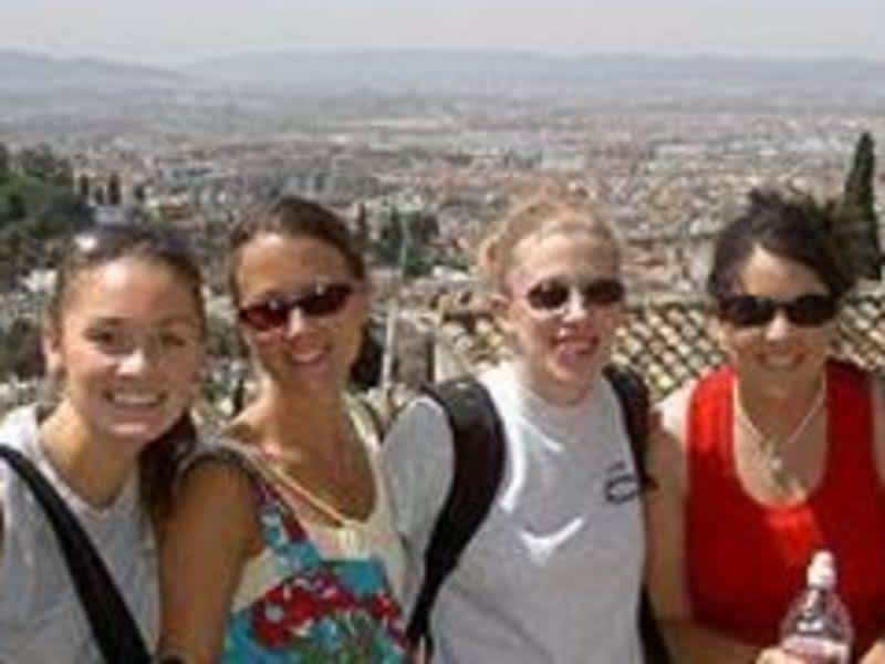 Four women smiling and posing for a picture on a hill, enjoying the beautiful scenery.