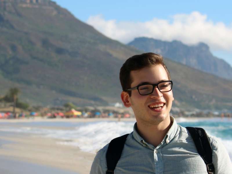 Male student smiles in front of Cape Town ocean and mountain backdrop