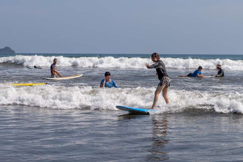 People surf in the waves, with a girl standing on board.
