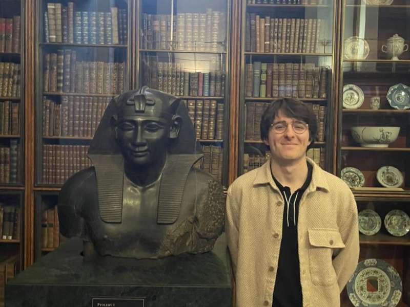 Person standing beside an ancient Egyptian statue in a library with bookshelves in the background.