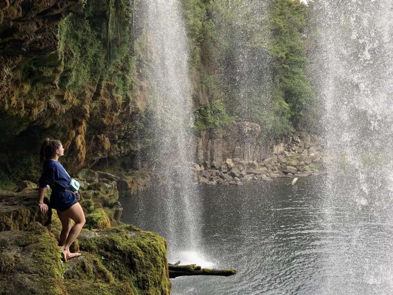 student standing beneath a waterfall and body of water