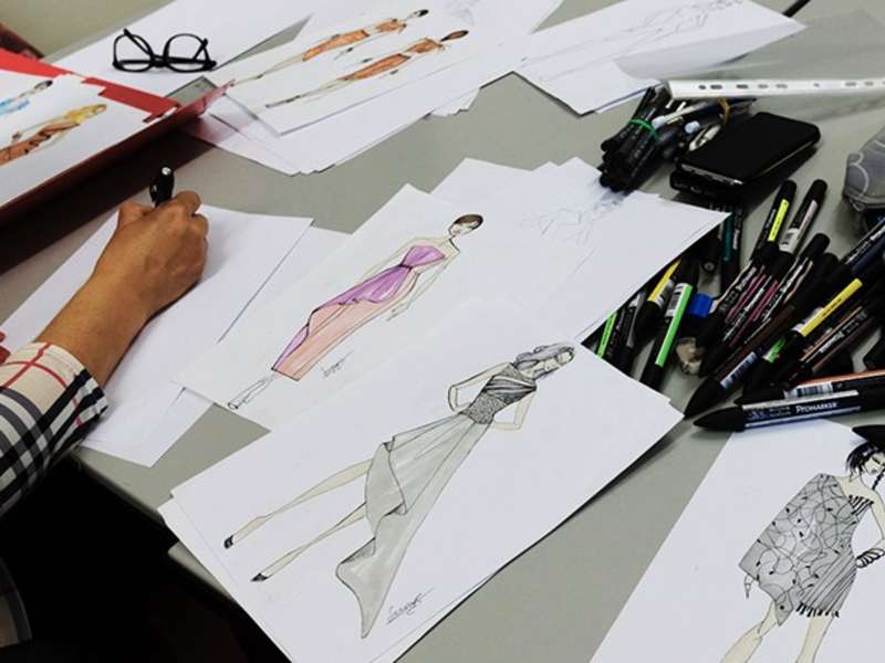 Student working on their fashion sketches, with a selection of sketches and pens on the table.