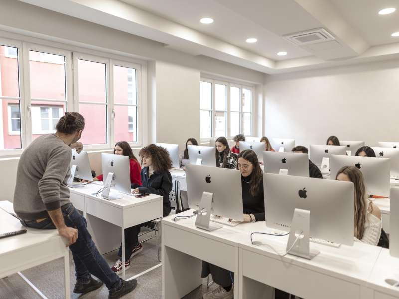 Students sat behind apple computers while a teacher sits on his desk in front of them