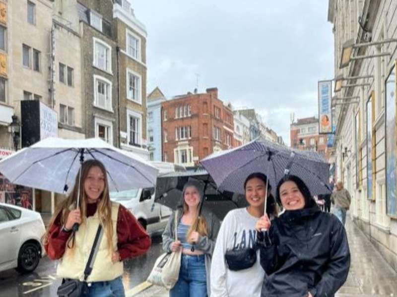 Students walk with umbrellas on a rainy street in London.