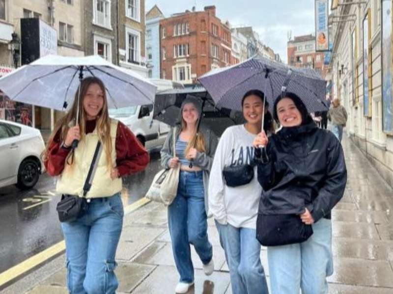 Students walk with umbrellas on a rainy street in London.