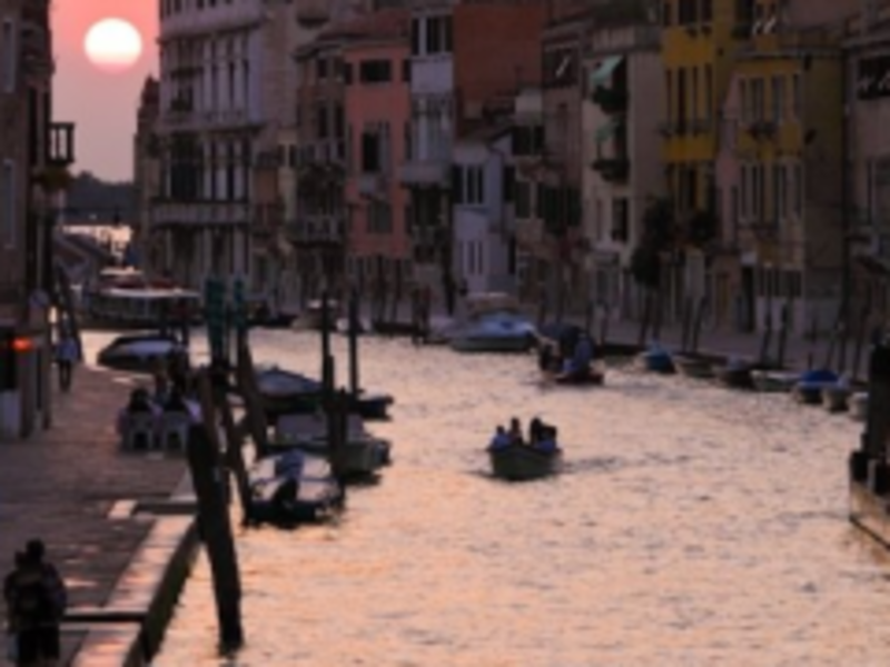 Sun setting over canal in Venice, casting golden hues on the water and historic buildings lining the banks.