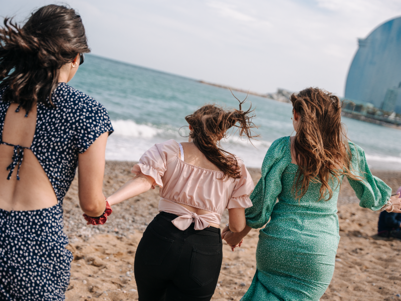 Three EF Gap Year students holding hands and running toward the waves on a sandy beach in Barcelona, with the iconic W Hotel visible in the background.