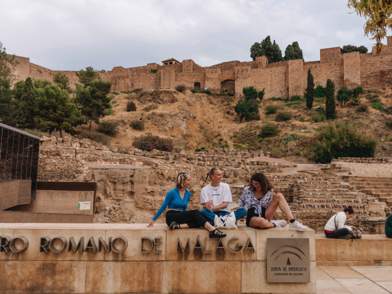 Three EF Gap Year students sitting by the Roman Theatre in Málaga with the Alcazaba fortress in the background, exploring Spain’s rich history and culture.