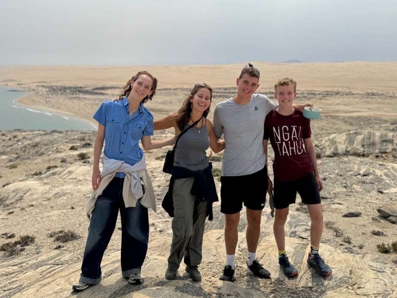 Three interns and a student pose together on a rocky clifftop overlooking a sweeping Namibian coastal landscape, with the desert plains and Atlantic Ocean stretching out behind them.