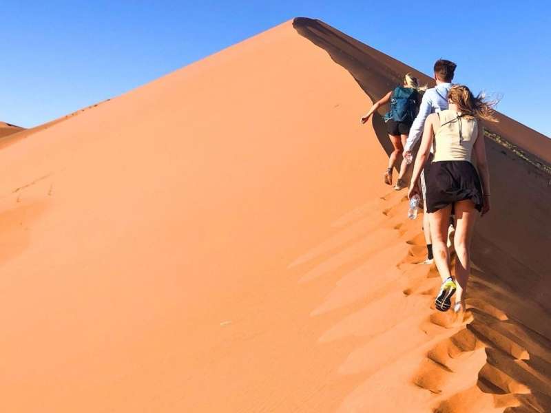 Three interns climb the steep face of a towering red sand dune at Sossusvlei, Namibia, under a clear blue sky.