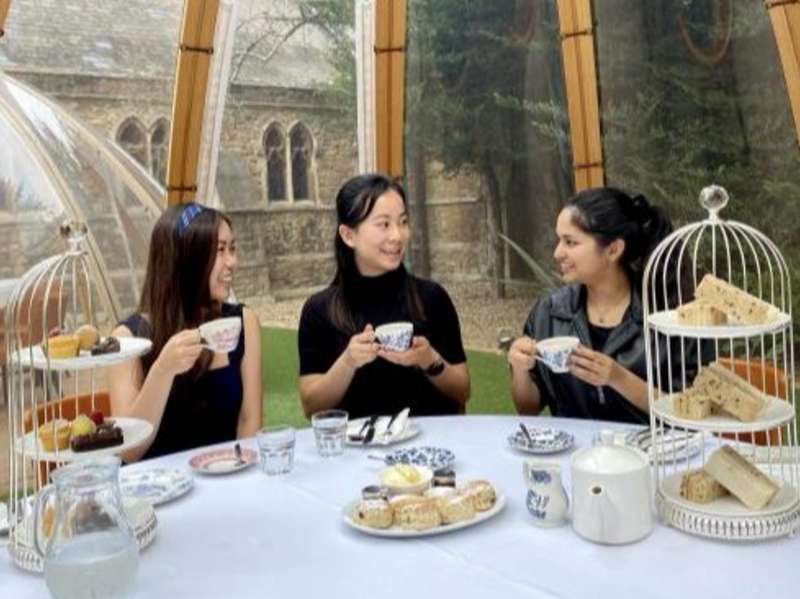 Three people seated at a table with tea cups, sandwiches, and pastries inside a glass conservatory.