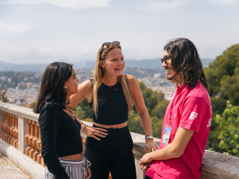 Three students talking and smiling on a scenic viewpoint in Nice, with the city and hills in the background.