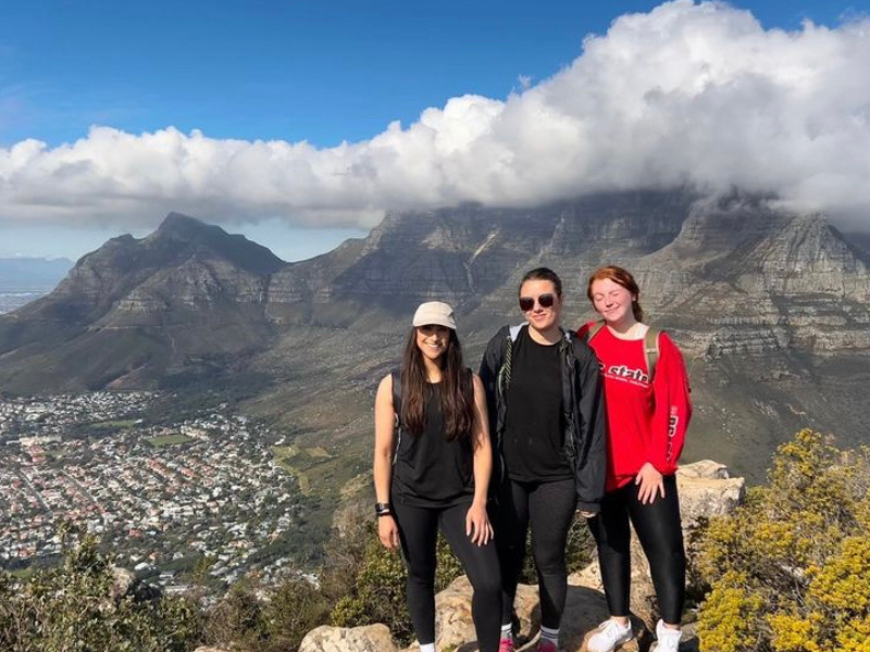 Three volunteers stand atop a mountain, with Cape Town in the background and the peak of table mountain shrouded in cloud. 