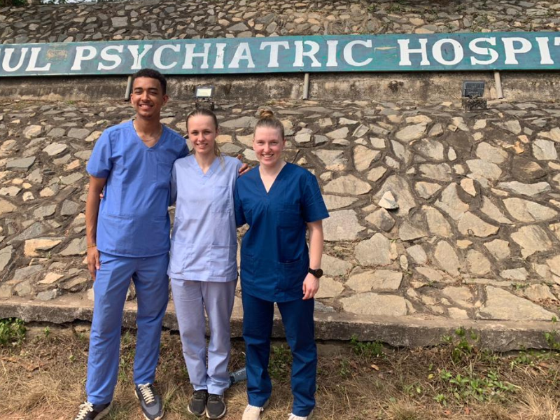 Three volunteers stand in their scrubs underneath a sign that reads 'psychiatric hospital'
