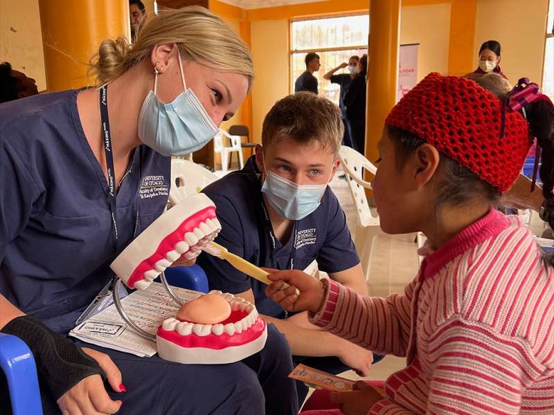 Two dental volunteers wearing masks and scrubs teach a young girl how to brush teeth using a large dental model in Cusco, Peru.