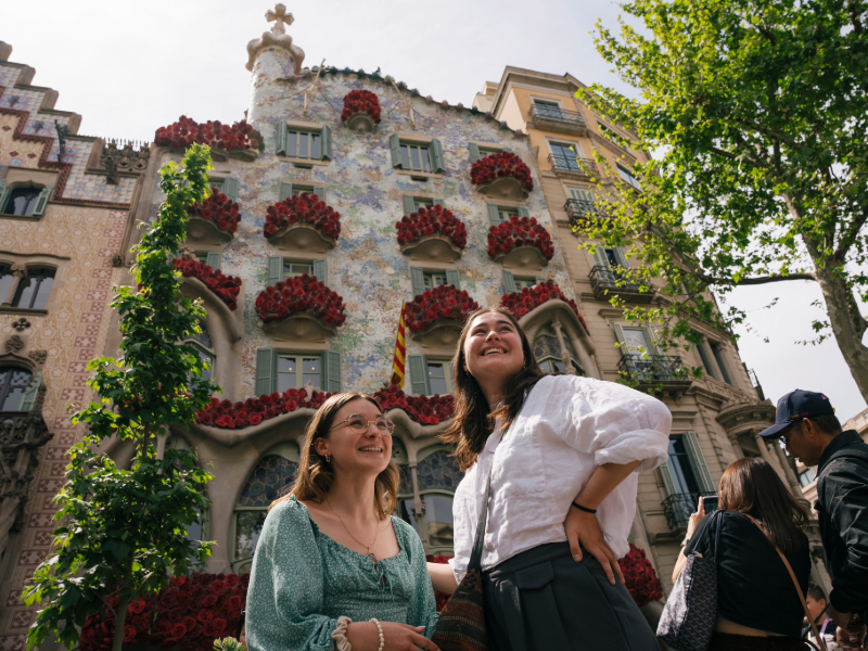 Two EF students smiling in front of Casa Batlló in Barcelona, decorated with red flowers on the balconies.