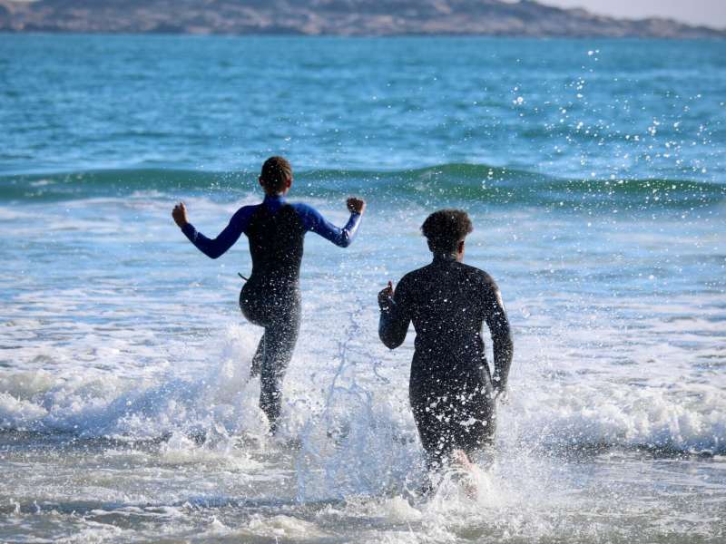 Two people in wetsuits run together into the waves of the South Atlantic Ocean on the Lüderitz coastline.