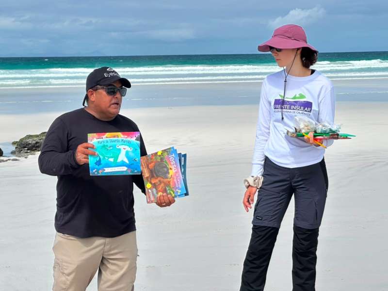 Two people stand on the beach, the man speaking with a stack of books.