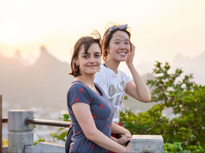Two smiling students lean on a stone railing at a hilltop viewpoint, with hazy karst peaks and a sunset sky behind them.