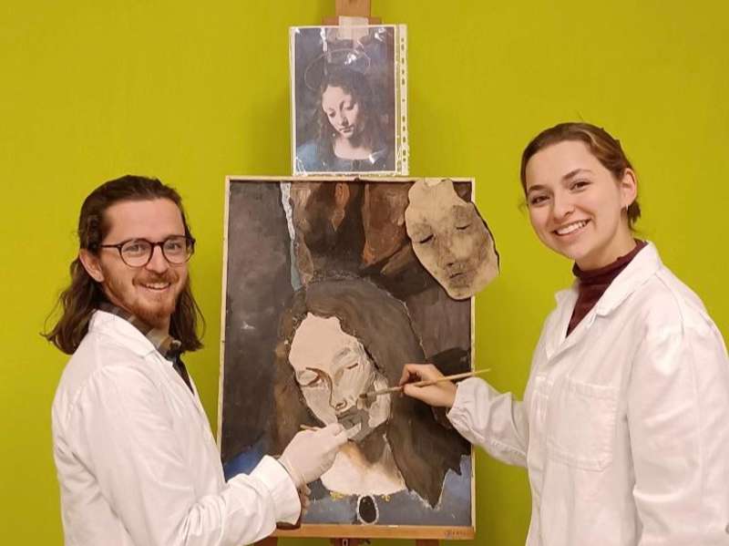 Two students in lab coats smiling in front of a fresco they are working on.