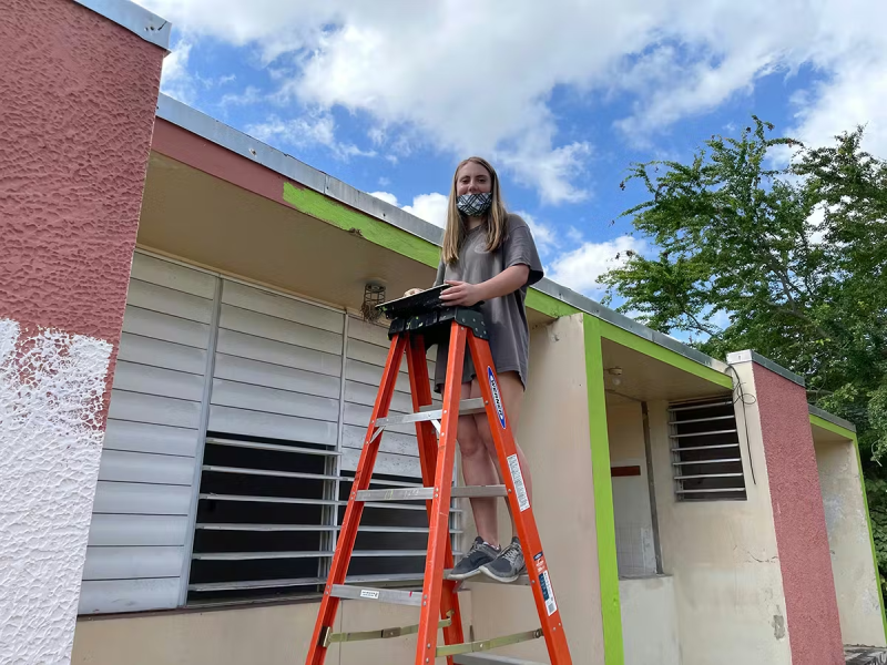 Volunteer standing on a ladder painting the trim of a school