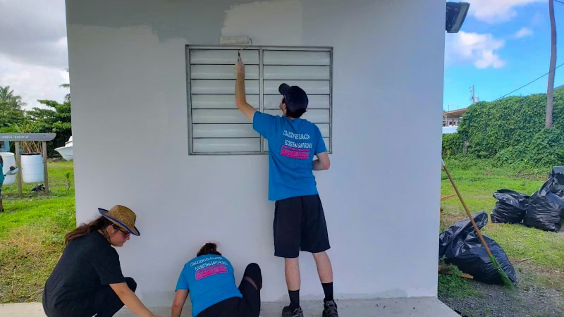 Volunteers painting the exterior wall of a small building with paint